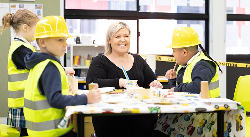 Dedicated teacher assisting Holy Family Primary Emerton students in class. The students are wearing hard hats and construction vests.